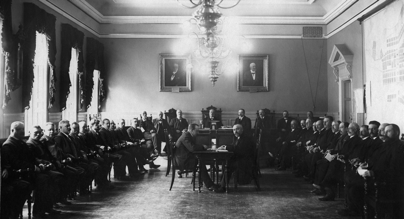Gruppbild med stadsfullmäktiges ledamöter som sitter på stolar placerade längs väggarna. Vid kortsidan ett bord och fler sittande ledamöter. I mitten ett bord med två personer som sitter mittemot varandra
