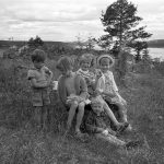 Tre barn sitter på en sten och i gräs med glas och muggar i händerna.