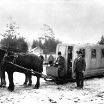 En kusk och två hästar som drar en överbyggd vagn. I vagnen finns flera människor som ser ut genom dörren eller fönster.