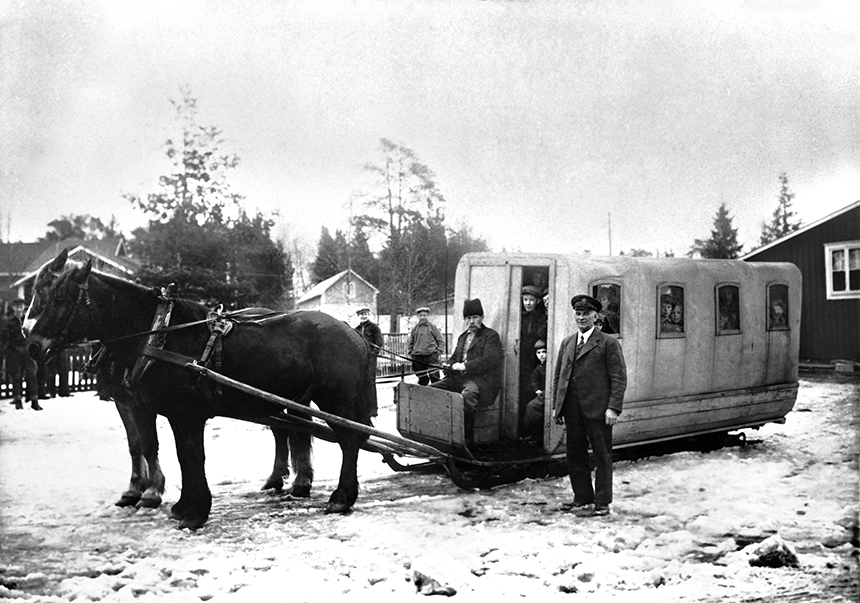 En kusk och två hästar som drar en överbyggd vagn. I vagnen finns flera människor som ser ut genom dörren eller fönster.