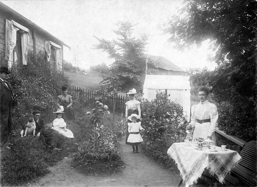 Dukat kaffebord i trädgård, en kvinna serverar kaffe och flera personer, både vuxna och barn och en hund finns på gräsmattan eller i trädgårdsgången