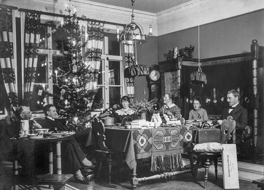 Bilden visar en familj som sitter vid ett bord. Ala är vuxna. I bakgrunden syns en julgran