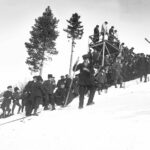 Bilden visar en snöig backe med domartorn i trä. Det är en tävling i backhoppning och i backen står många människor och väntar.