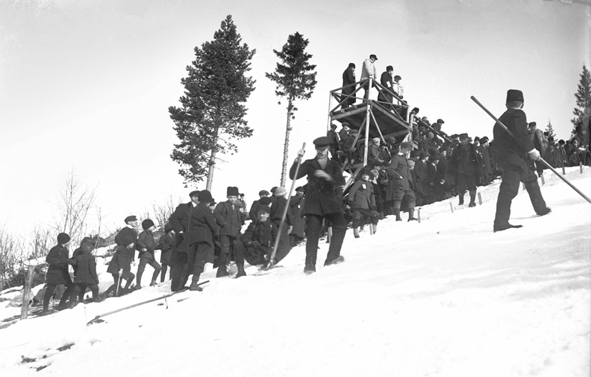 Bilden visar en snöig backe med domartorn i trä. Det är en tävling i backhoppning och i backen står många människor och väntar.
