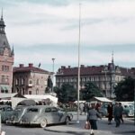 Bilden visar ett torg fyllt med torgstånd, människor och parkerade bilar