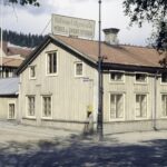 Bilden visar ett gammalt trähus med en gul brevlåda på gaveln. I bakgrunden skymtas skog.
