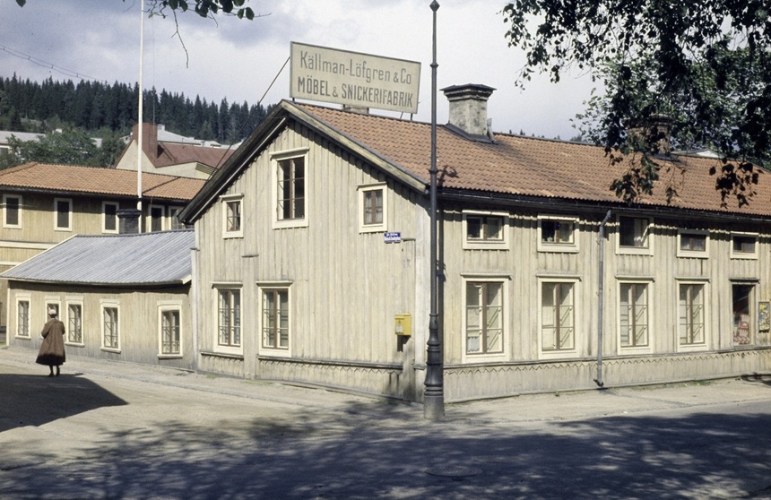 Bilden visar ett gammalt trähus med en gul brevlåda på gaveln. I bakgrunden skymtas skog.